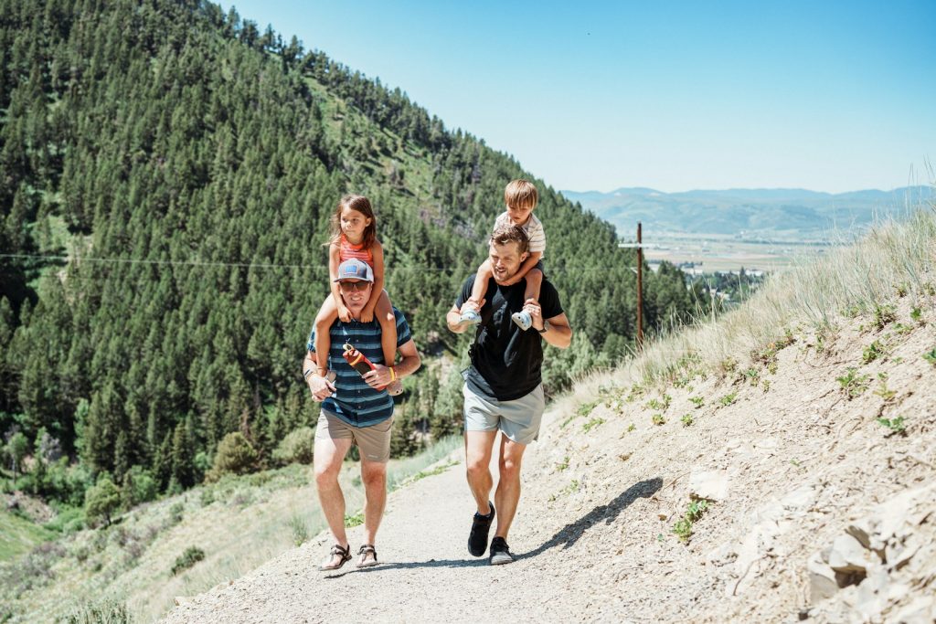 Two fathers carrying children on shoulders on a mountain path.
